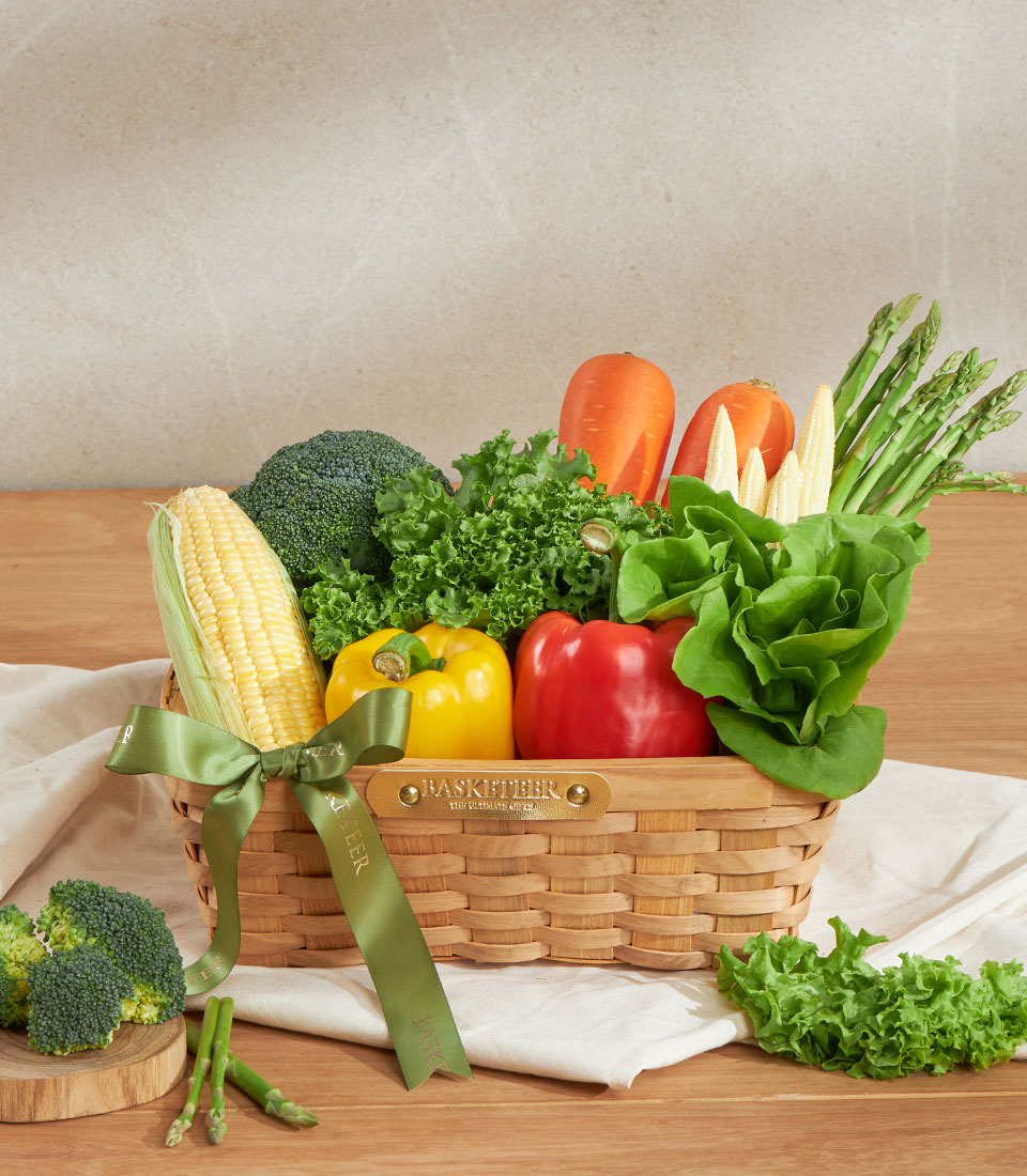 Woven wooden basket of fresh vegetables with corn, broccoli, peppers, and leafy greens, tied with sage green ribbon