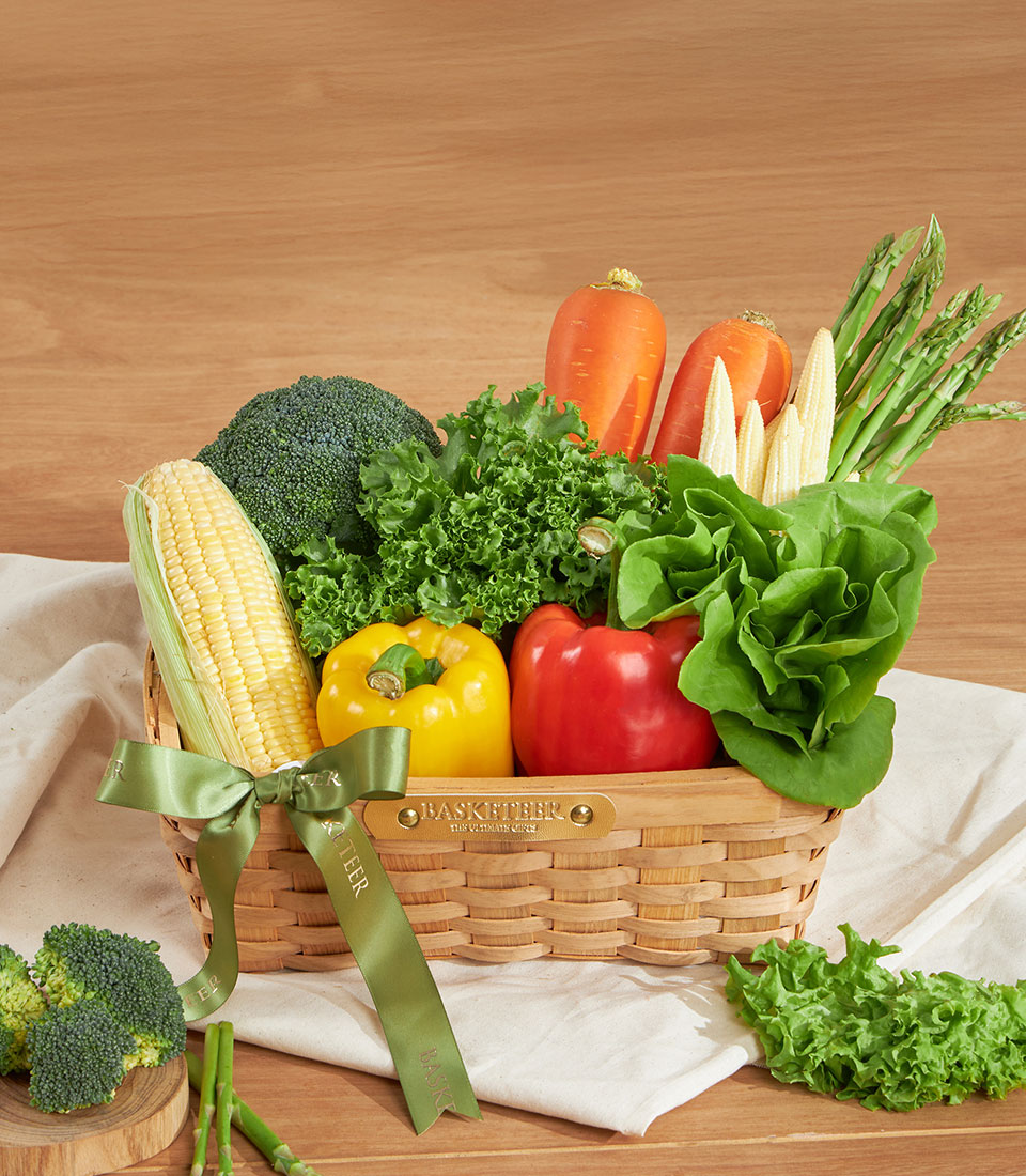 Wooden basket filled with fresh vegetables including corn, broccoli, bell peppers, carrots, and leafy greens with green