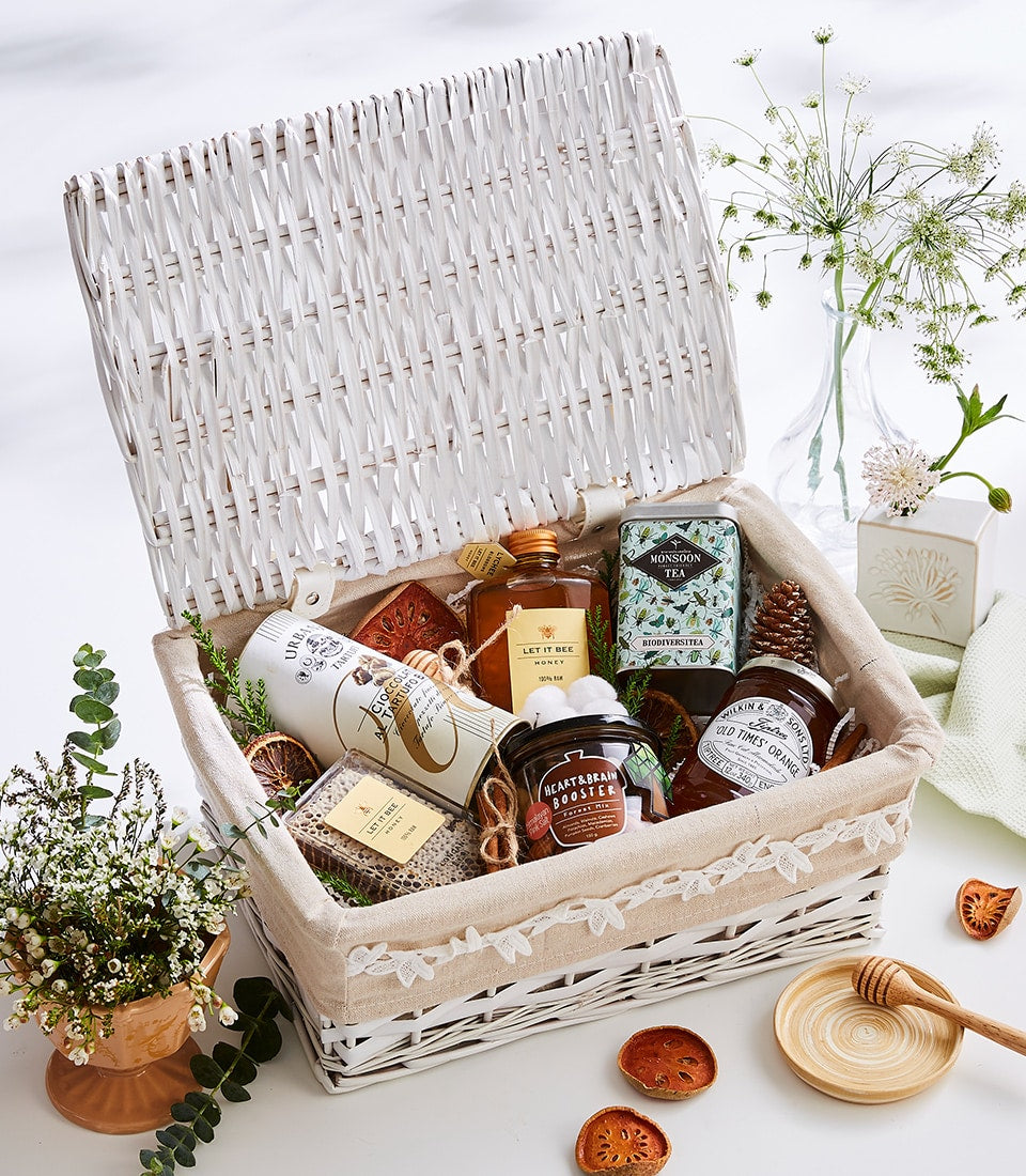 Curated white wicker gourmet gift basket with honey, tea, preserves, and artisan chocolates, styled with dried citrus and