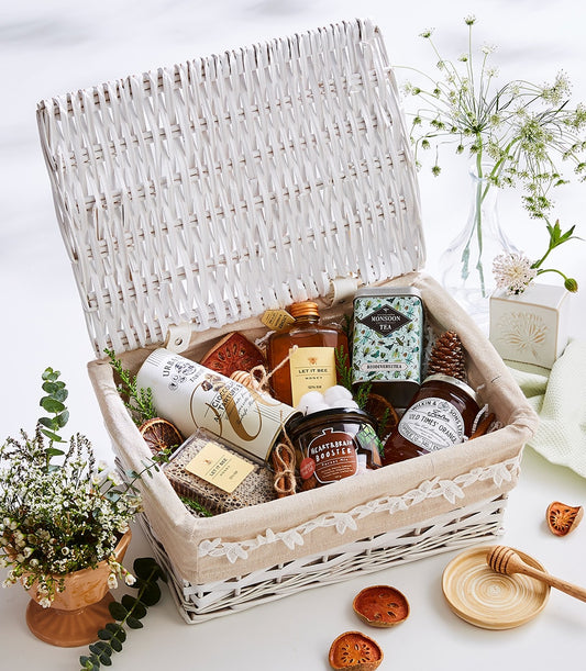 Curated white wicker gourmet gift basket with honey, tea, preserves, and artisan chocolates, styled with dried citrus and