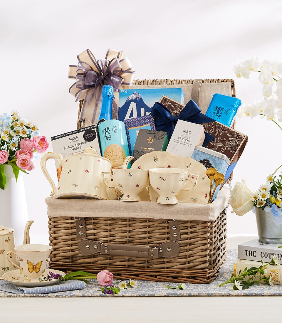 Wicker picnic basket with gourmet treats, floral teacup set, and grey-purple ribbon bow