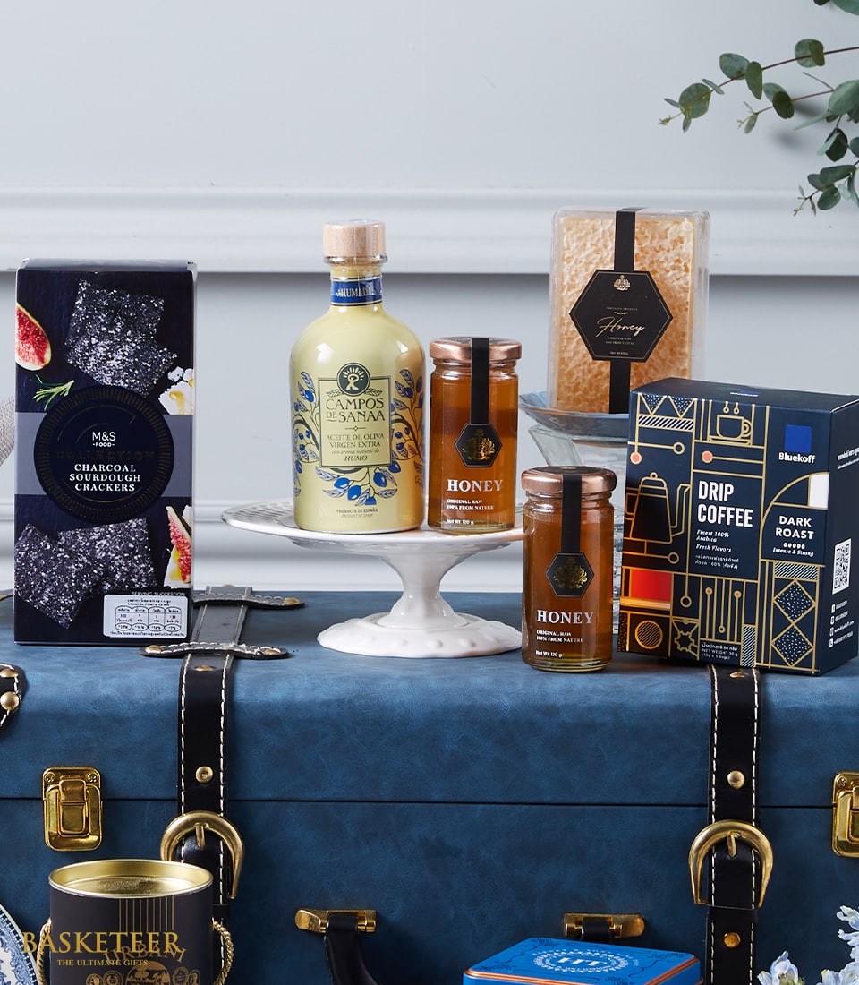 Luxury gourmet gift basket with olive oil, honey, honeycomb, sourdough crackers, and drip coffee on vintage blue trunk
