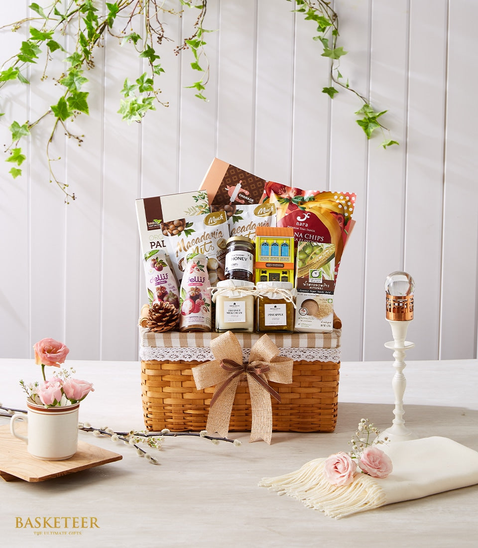 Woven wicker gift basket with gourmet Thai snacks, honey, and coconut products, styled with pink roses and cream candleholder