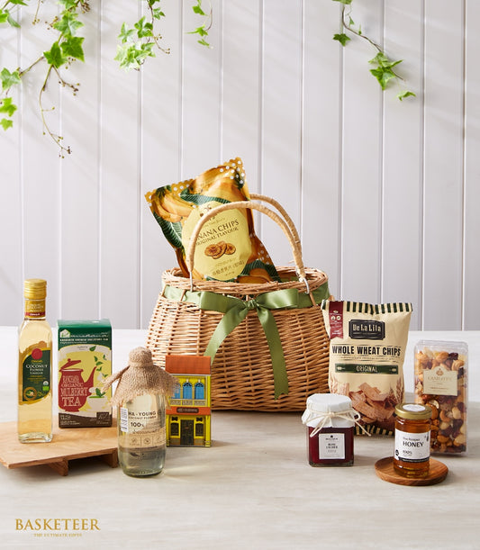 Woven gift basket with green ribbon filled with Thai gourmet treats including coconut flower vinegar, mulberry tea, honey,
