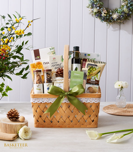 Woven gift basket with green ribbon containing gourmet wellness items, olive oil, coconut waffles, dried mango, and spice
