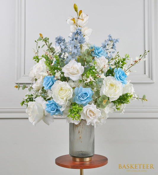 Light blue and white artificial flower arrangement in ribbed glass vase with gold base and wooden stand