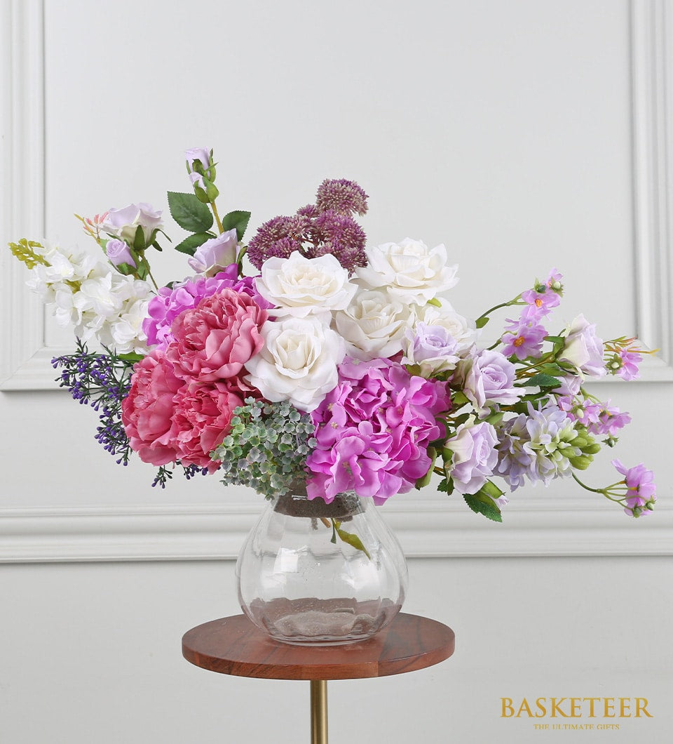 Purple and pink artificial flowers in clear glass vase with peonies, roses, and hydrangeas