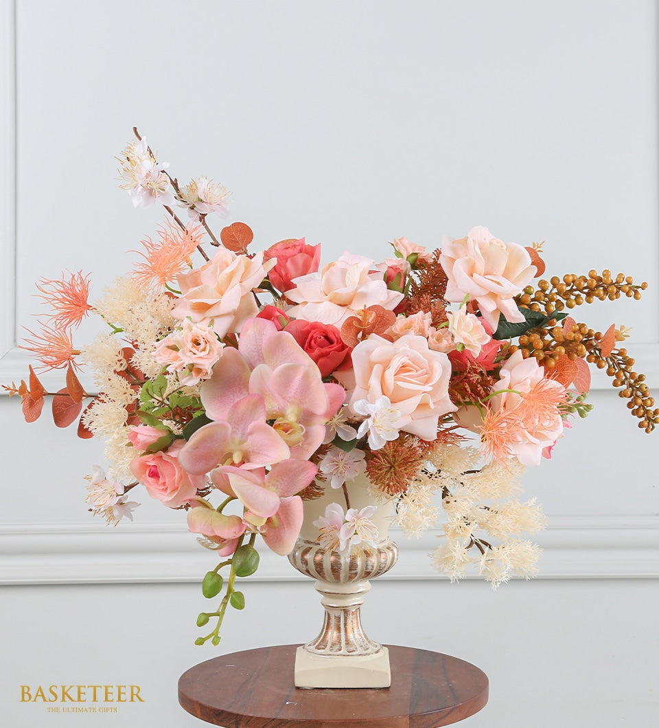 Peach and coral artificial flower arrangement in cream pedestal vase with gold accents, featuring roses and orchids
