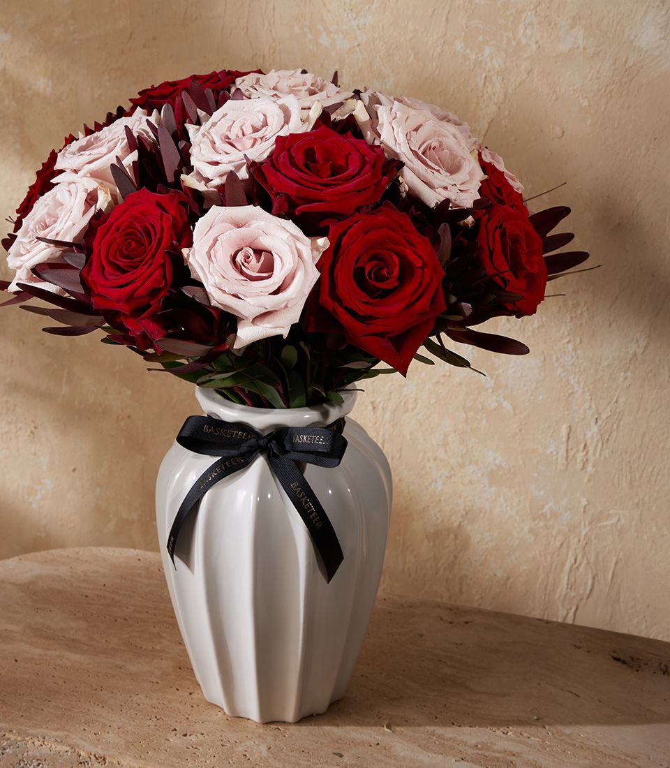 Red and light pink roses arranged in white ceramic vase with black ribbon bow, dark foliage, premium fresh flower gift