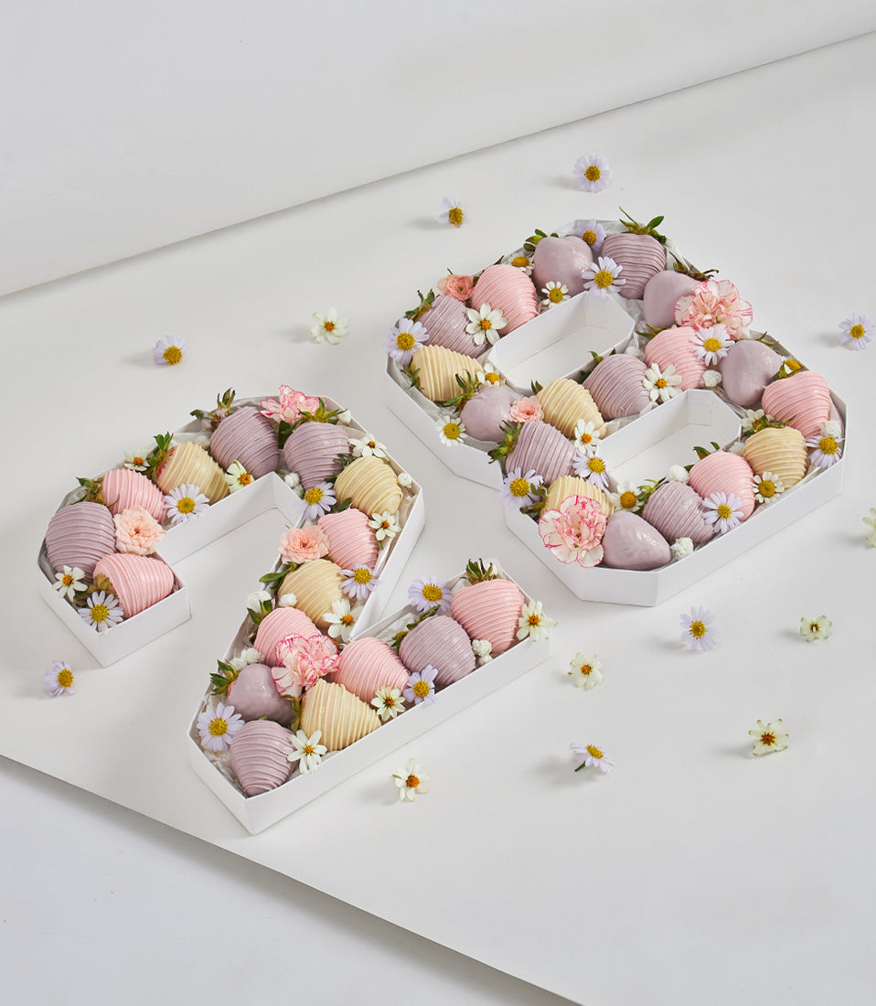 Chocolate-dipped strawberry number box "29" with pastel pink, yellow, and lavender coating, fresh flowers, and foliage for