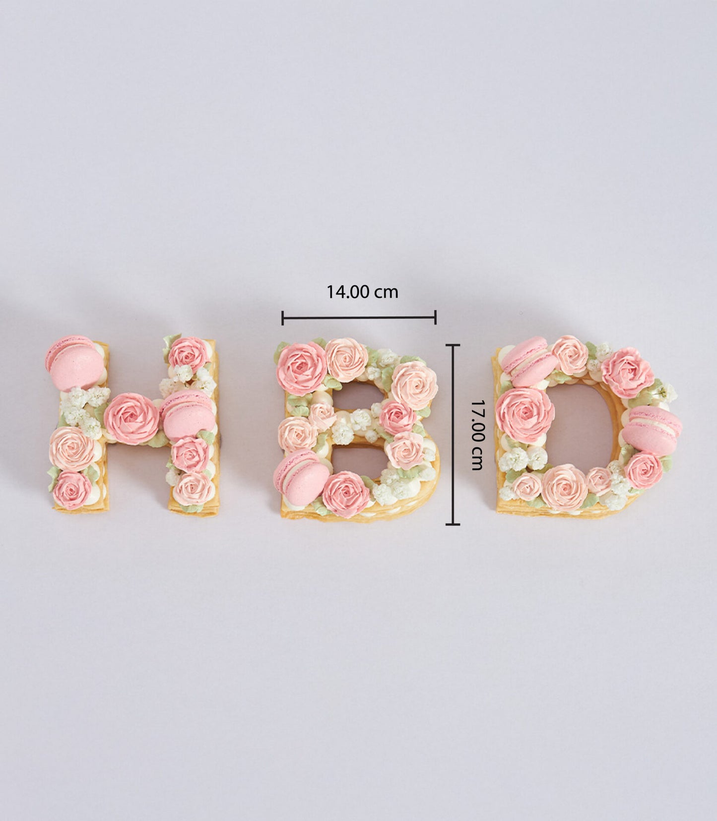 Cream pastry letter cakes spelling HBD, decorated with pink roses, macarons, and white flowers for birthday celebrations