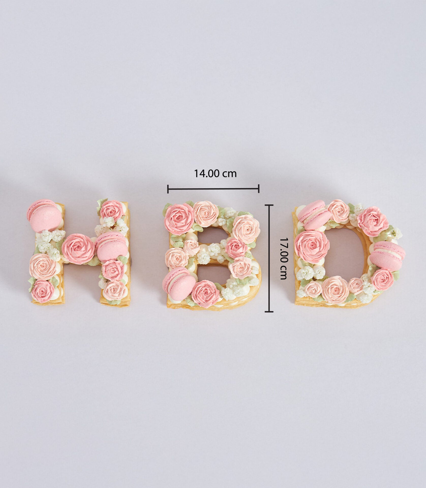 Cream pastry letter cakes spelling HBD, decorated with pink roses, macarons, and white flowers for birthday celebrations