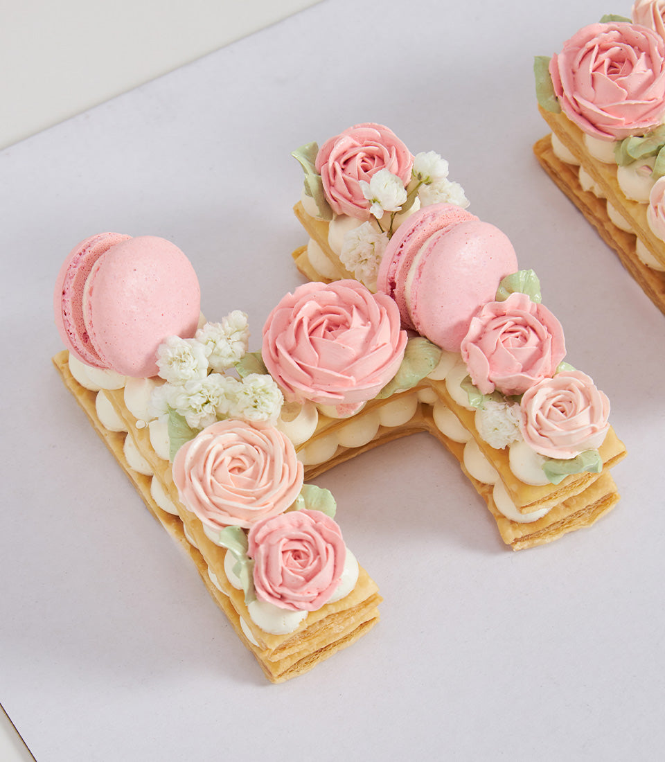 Monogram letter H pastry cake with cream layers, pink buttercream roses, and macarons for birthday celebrations