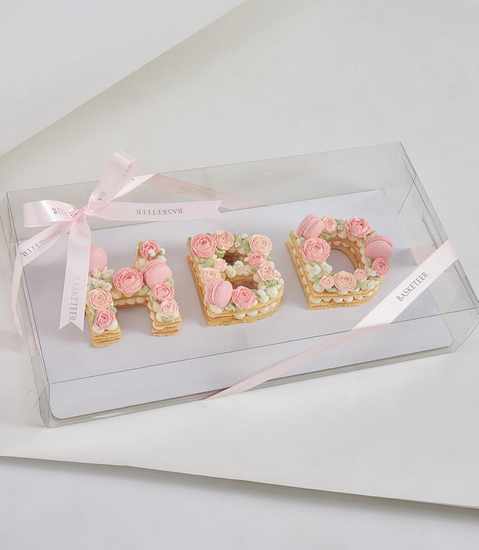 Letter-shaped cream tarts spelling HAB with white cream, pink piped roses, and macarons in luxury gift box with pink ribbon