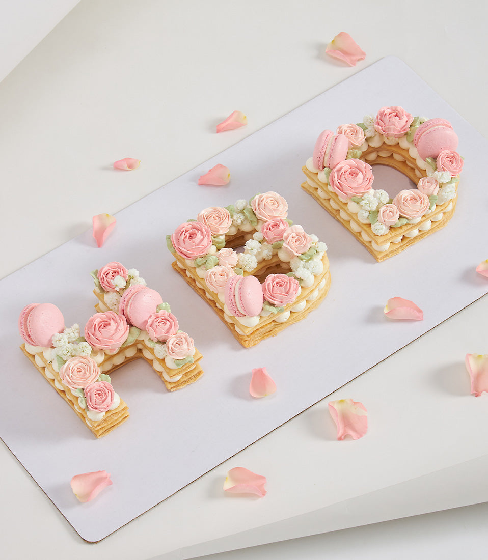 Golden cream letter tarts spelling HBD with pink roses, macarons, and rose petals for birthday celebration