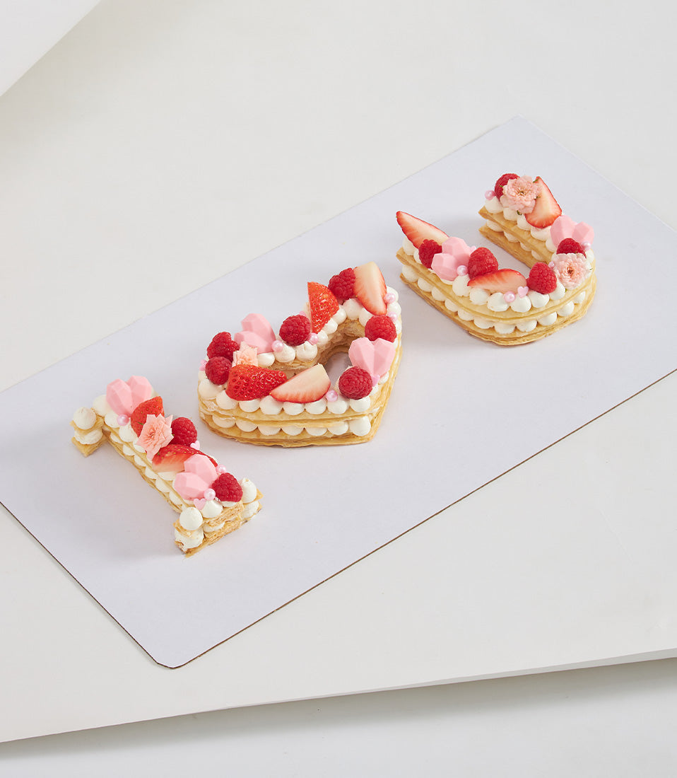Three letter cakes spelling "I ❤️ U" with strawberries, raspberries, and cream on white board