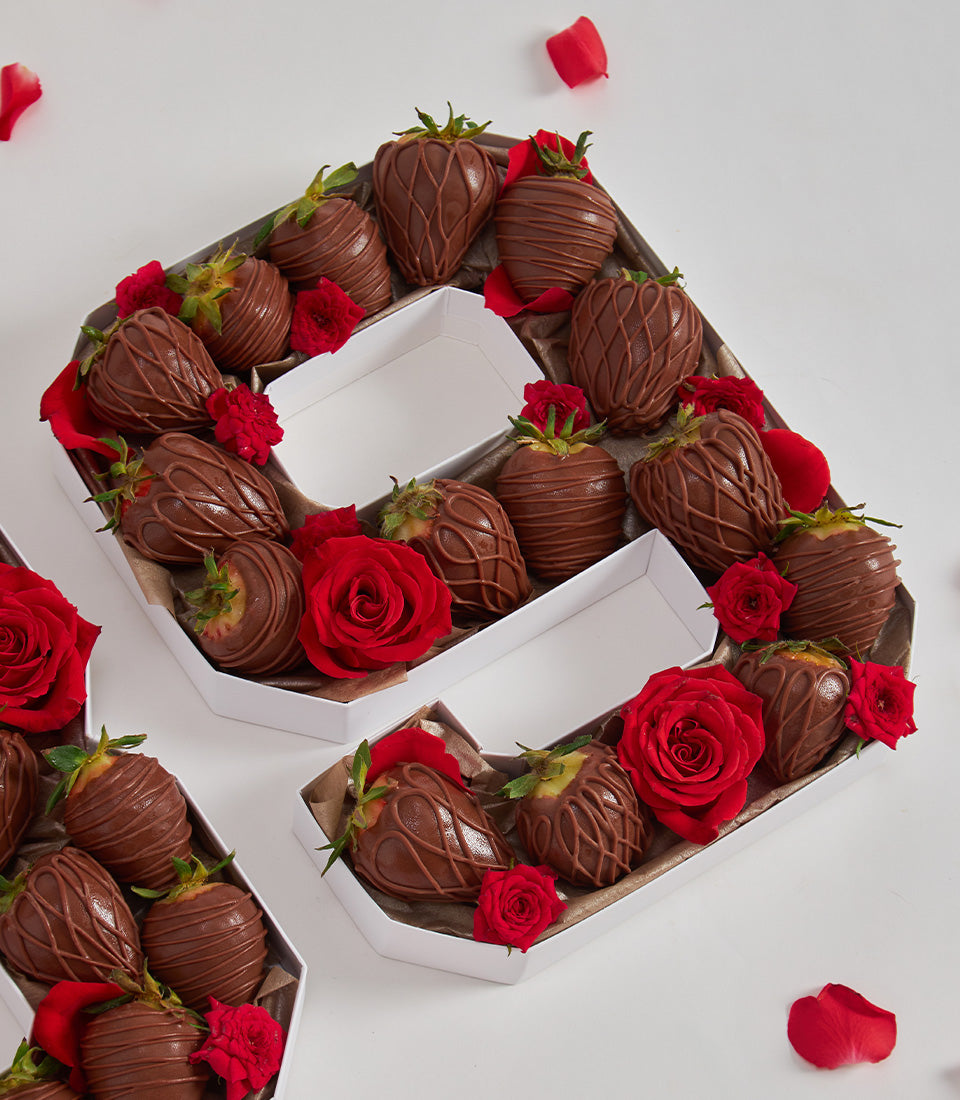 Chocolate-covered strawberries arranged in letter boxes spelling LOVE with red roses and petals