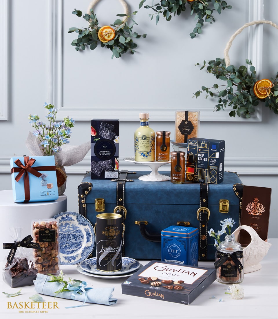 Luxury gift hamper with gourmet foods, chocolates, and blue flowers arranged on dark leather trunk with gold accents
