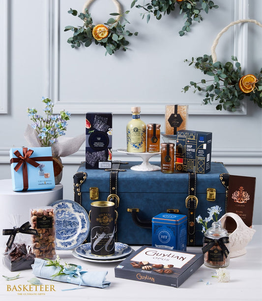 Luxury gift hamper with gourmet foods, chocolates, and blue flowers arranged on dark leather trunk with gold accents