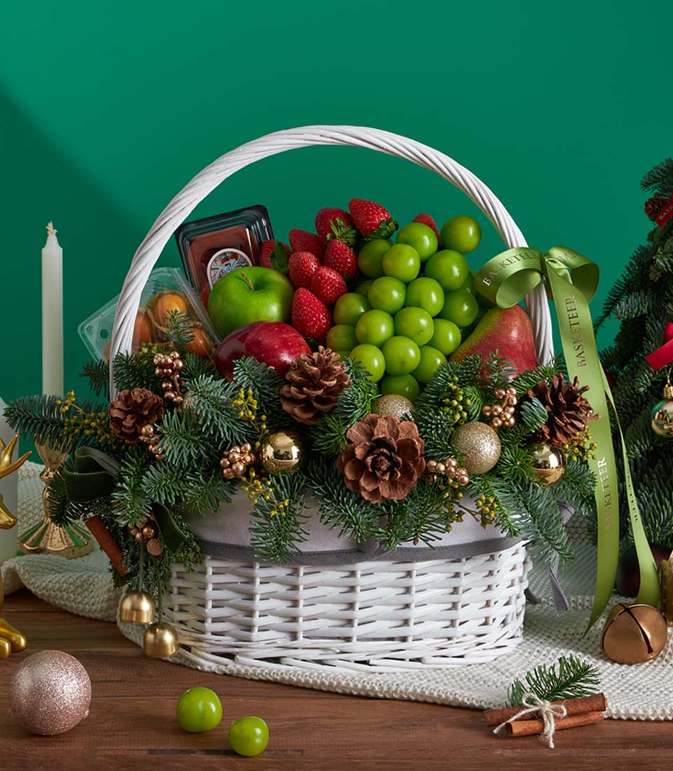 White wicker Christmas fruit basket with fresh strawberries, grapes, apples, and mandarins, decorated with pine branches and