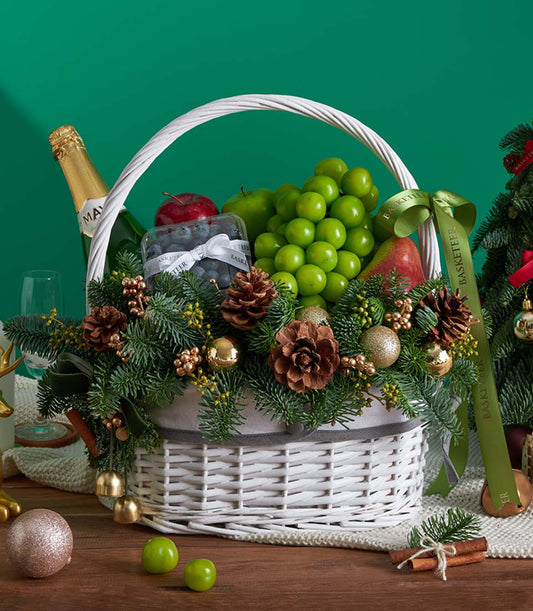 Festive white wicker Christmas fruit basket with apples, grapes, blueberries, pear, sparkling wine, pine branches, and gold