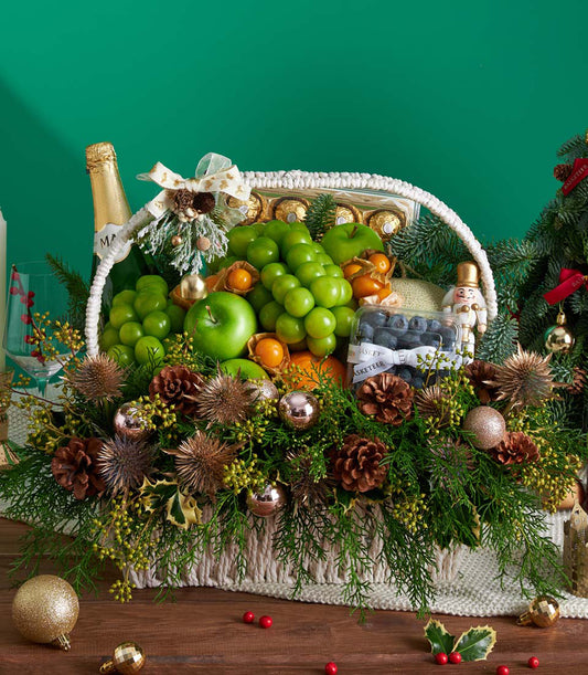 Festive white woven basket with fresh grapes, apples, blueberries, chocolates, and sparkling wine, decorated with gold