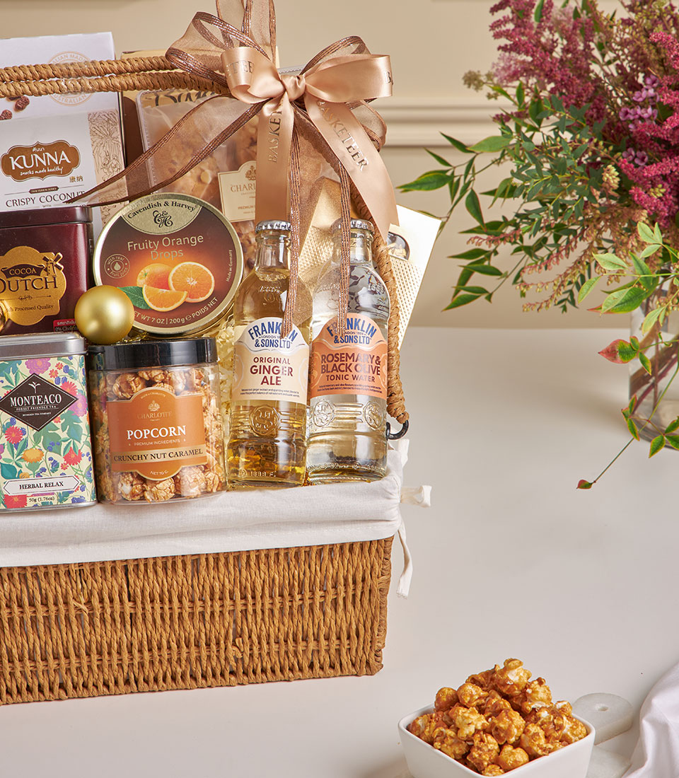 Woven wicker gift basket with gourmet snacks, herbal tea, artisan cookies, and tonic waters in warm earth tones for New Year
