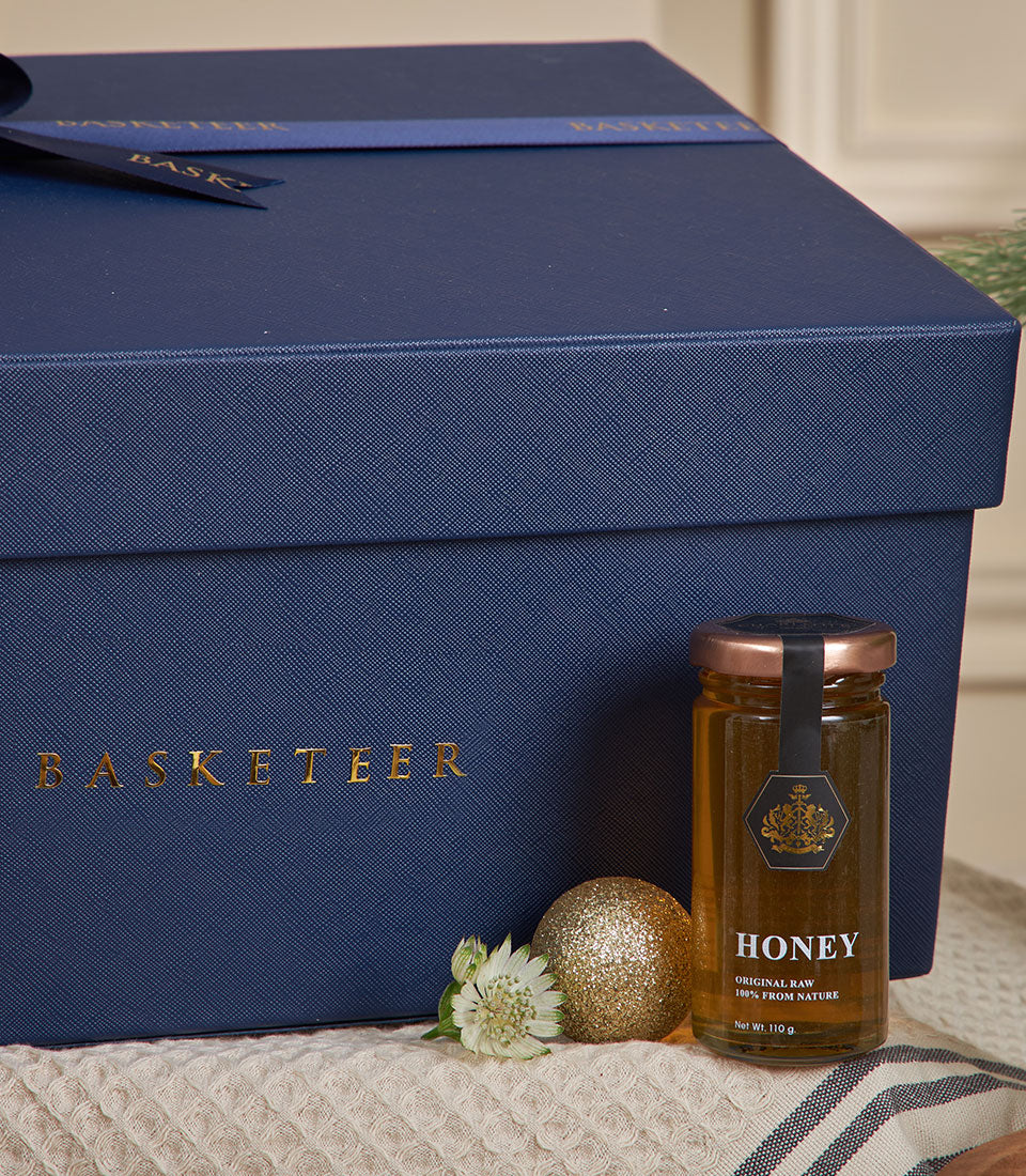 Dark blue luxury gift box with gold Basketeer ribbon, paired with raw honey jar and white flower for New Year gifting