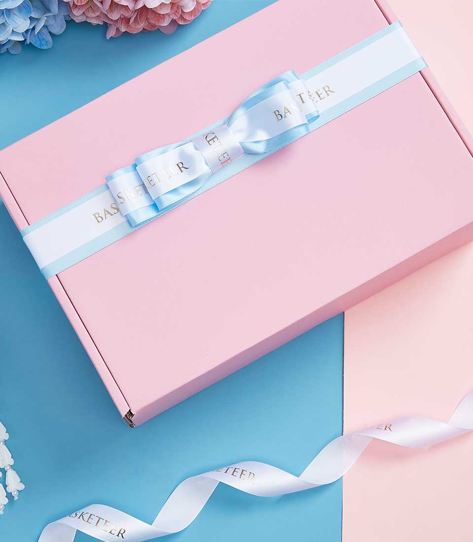 Light pink Mother's Day gift box with white ribbon bow and gold Basketeer branding, paired with fresh blush flowers