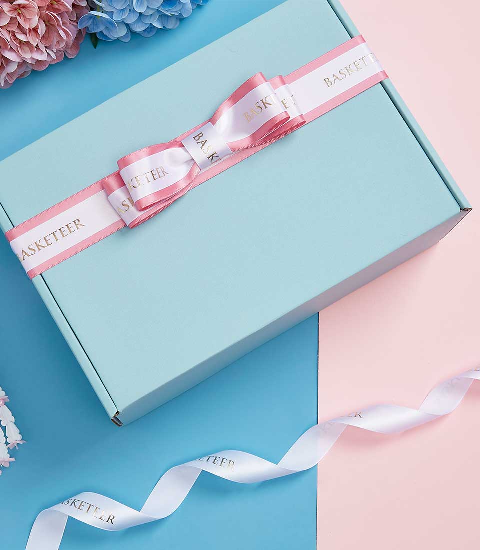 Light blue Mother's Day gift box with white ribbon bow and gold Basketeer lettering, styled with pink hydrangeas
