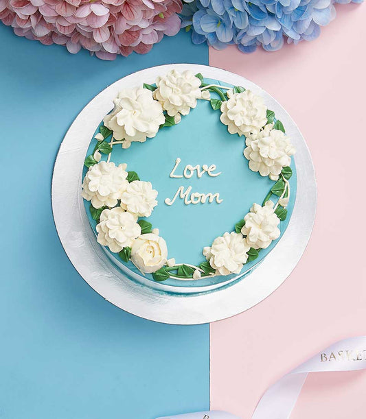Light blue birthday cake with white buttercream flowers and "Love Mom" inscription, presented with fresh floral arrangements