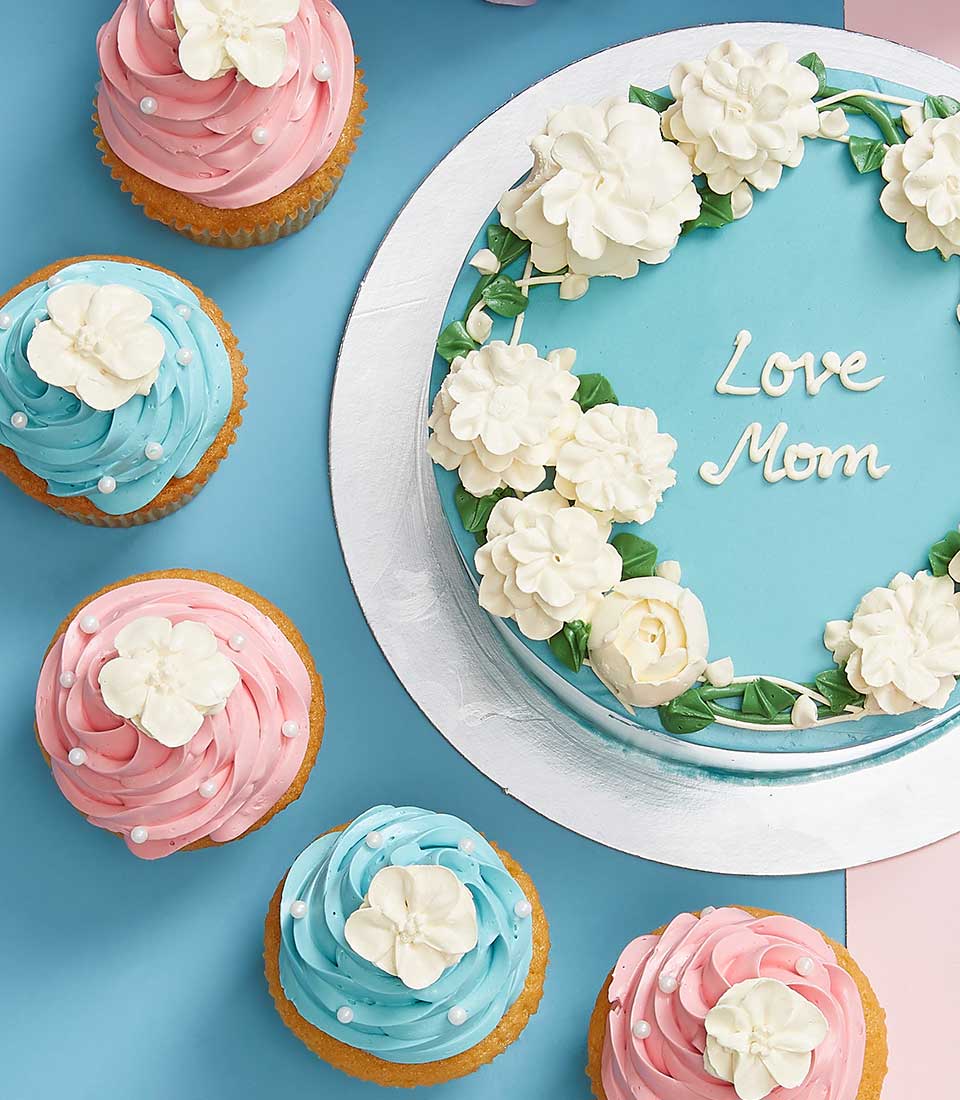 Light blue Mother's Day cake with white buttercream flowers and six matching cupcakes in pink and blue frosting