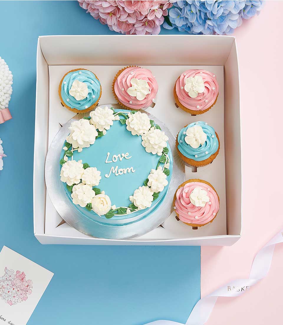 Light blue cake with white piped flowers and "Love Mom" message, surrounded by blue and pink frosted cupcakes with pearl