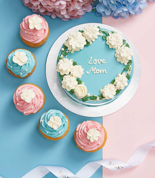 Light blue Mother's Day cake with white floral wreath and six pastel cupcakes in pink and blue frosting, topped with white