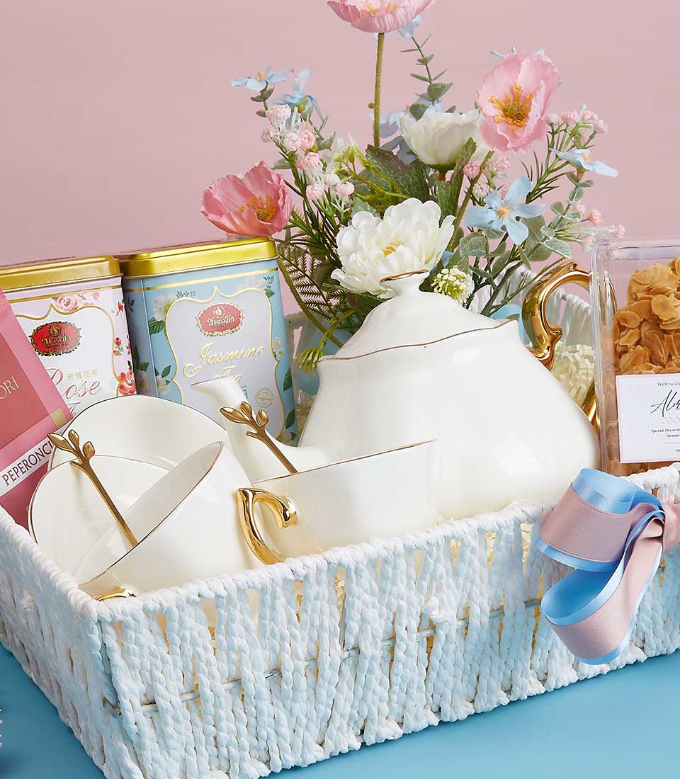 White woven gift basket with ceramic tea set, rose and jasmine tea tins, almond cookies, and pink silk flowers for Mother's