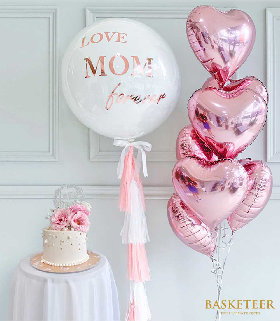Elegant white Mother's Day cake with pink roses and gold accents, paired with rose gold "LOVE MOM" balloon and pink heart