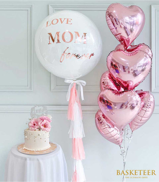 Elegant white Mother's Day cake with pink roses and gold accents, paired with rose gold "LOVE MOM" balloon and pink heart
