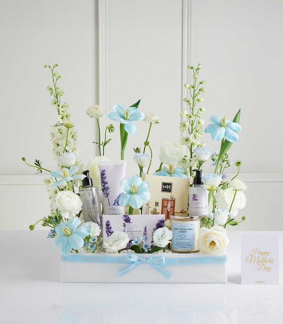 Luxury spa gift basket with lavender lotion, soap, and linen-scented candle surrounded by white delphiniums and light blue