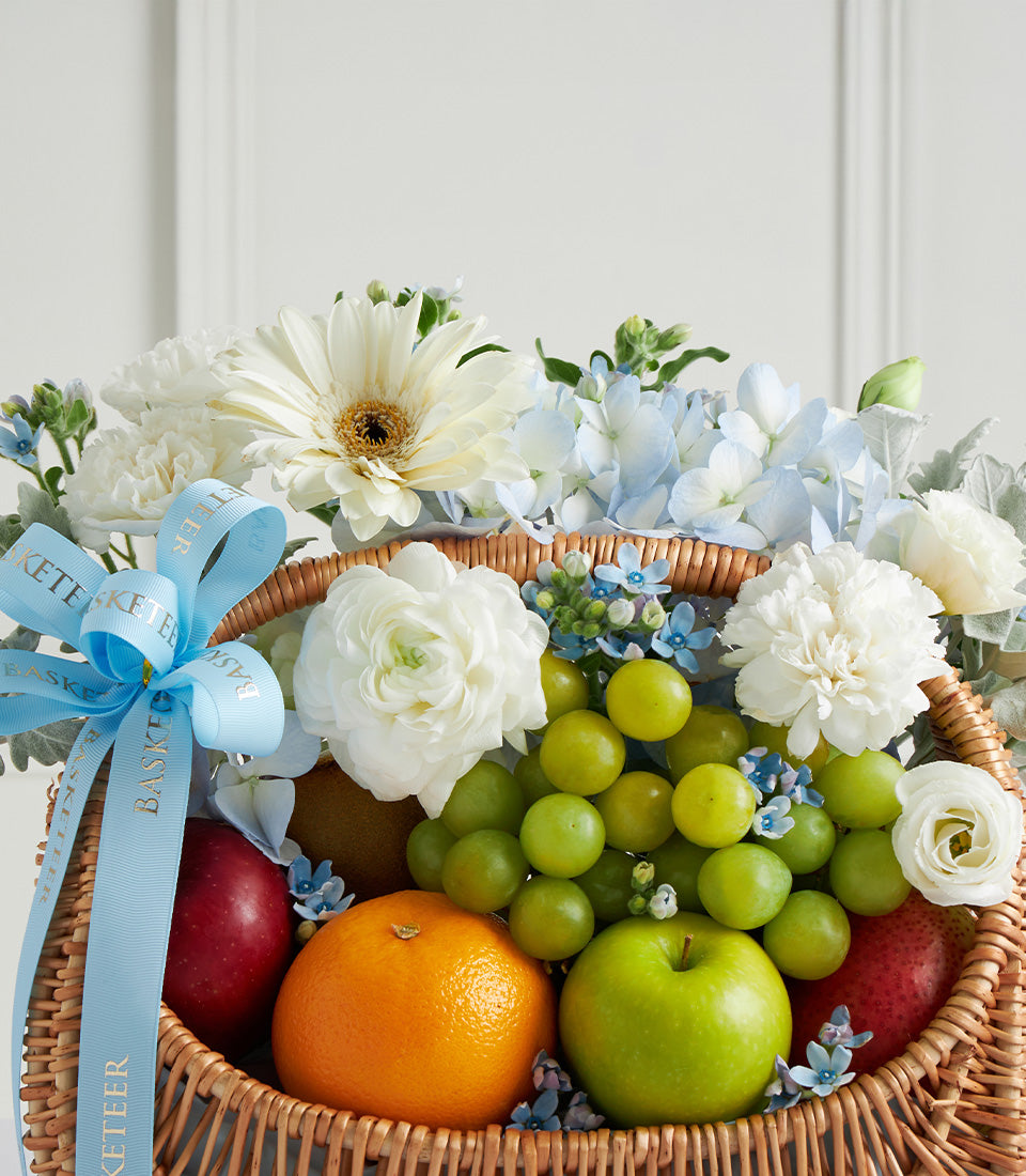 Wicker fruit and flower gift basket with fresh apples, oranges, grapes, white gerberas, ranunculus, and blue hydrangeas for