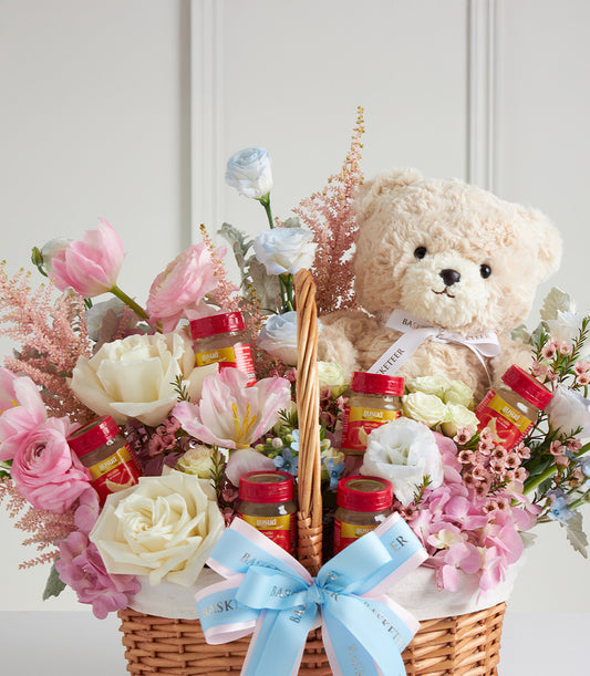 Light brown teddy bear nestled in fresh roses, ranunculus, and tulips with woven basket and blue satin bow for Mother's Day