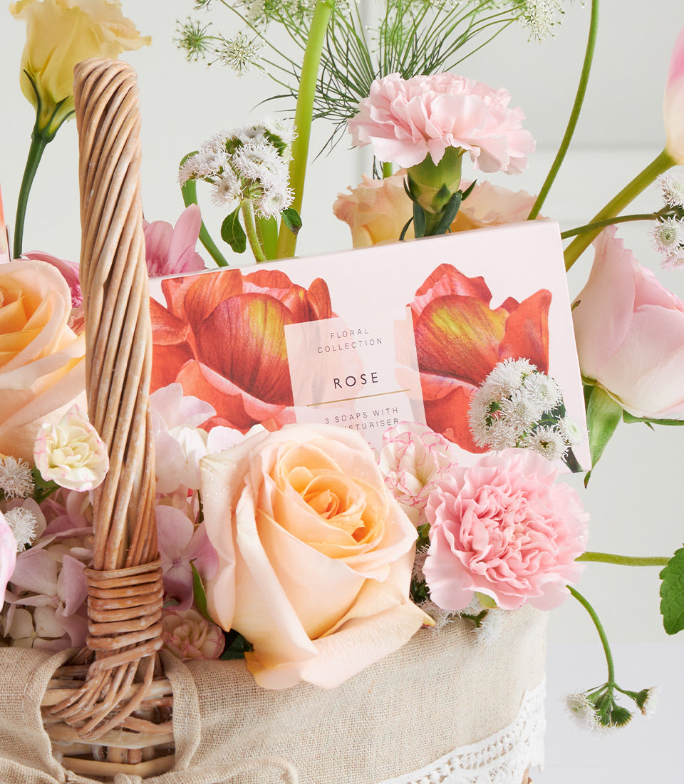 Luxury rose soap set with moisturiser in wicker basket, surrounded by fresh peach roses and pink carnations for Mother's Day
