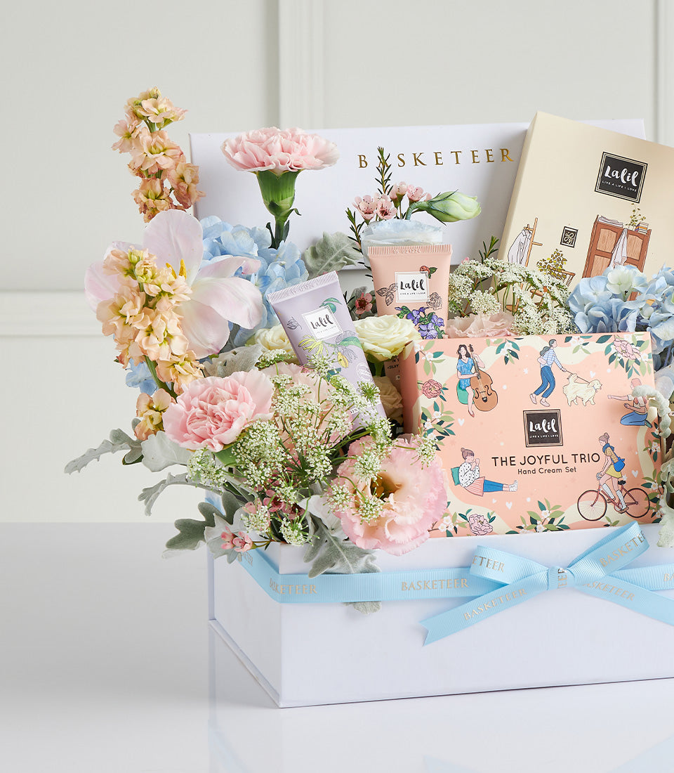 Luxury Mother's Day gift box with fresh pink and peach flowers, hand cream tubes, and beauty products tied with blue ribbon