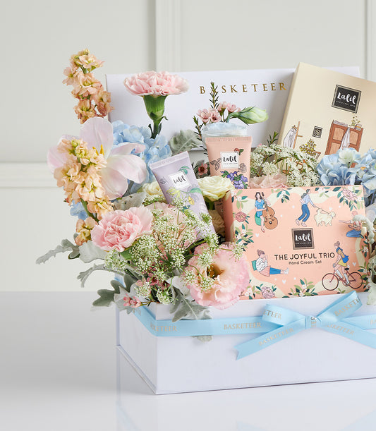 Luxury Mother's Day gift box with fresh pink and peach flowers, hand cream tubes, and beauty products tied with blue ribbon