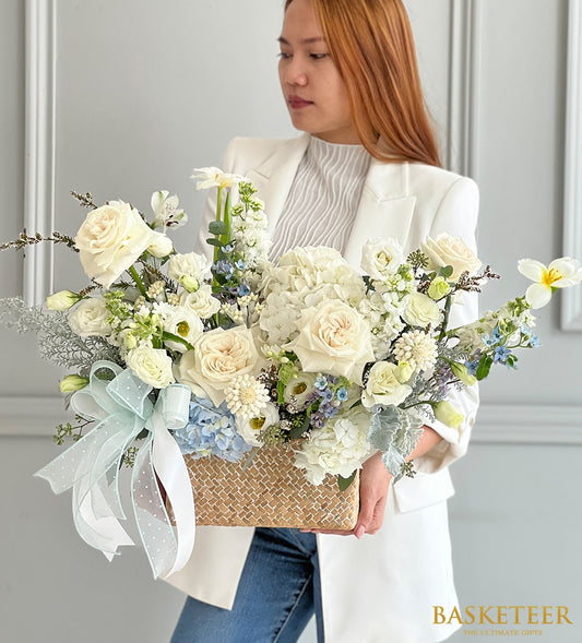 Large woven basket of white roses, hydrangeas, and cream flowers with blue accents, tied with polka-dot ribbon for get well