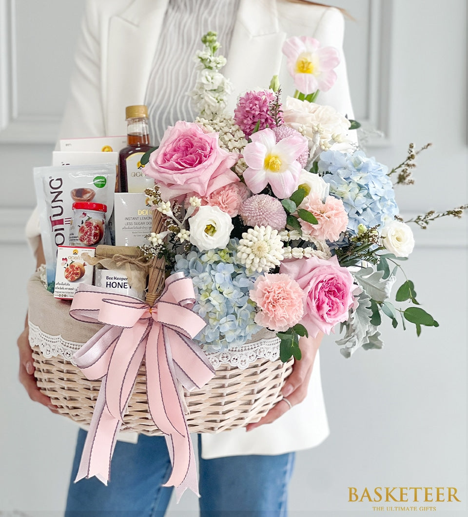 Luxury wicker gift basket with fresh pink and white flowers, Doi Tung and Doi Kham gourmet items, tied with pink ribbon