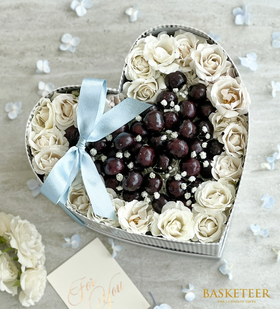 Heart-shaped gift box with dark cherry truffles, white roses, and baby's breath flowers tied with light blue ribbon