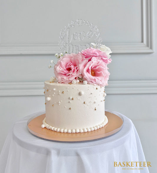 Mini white celebration cake with pink roses, white filler flowers, and gold pearl details on clear acrylic board