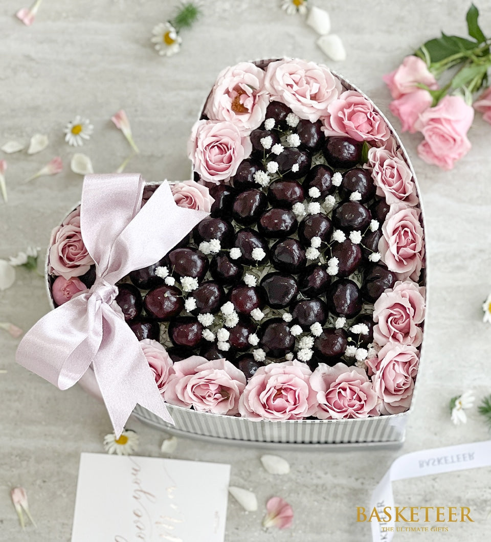 Heart-shaped gift box with dark cherries, pink roses, and baby's breath flowers tied with pink satin ribbon
