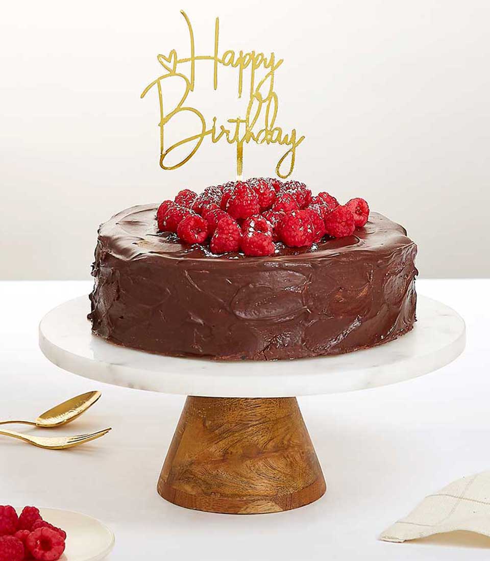 Decadent chocolate cake with rich ganache, fresh raspberries, and golden Happy Birthday topper on marble stand