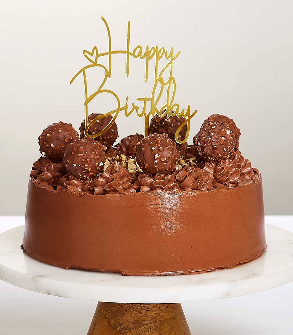 Round chocolate birthday cake with Ferrero Rocher chocolates, chocolate rosettes, and golden Happy Birthday topper on marble